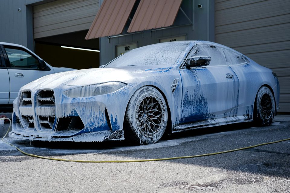 A car covered in thick wash foam being sprayed in a wash bay
